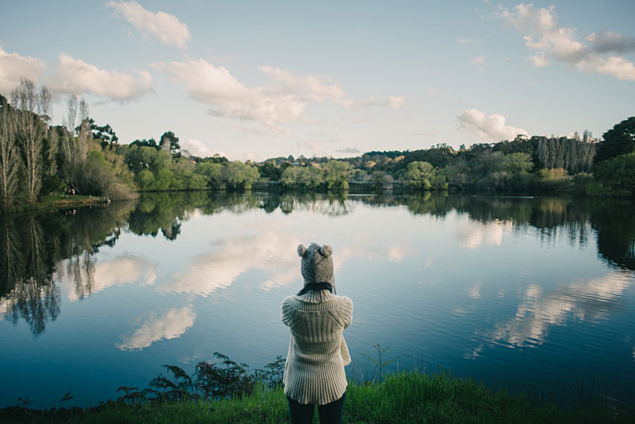 Child in a knitted hat standing at the edge of a still lake reflecting clouds and trees