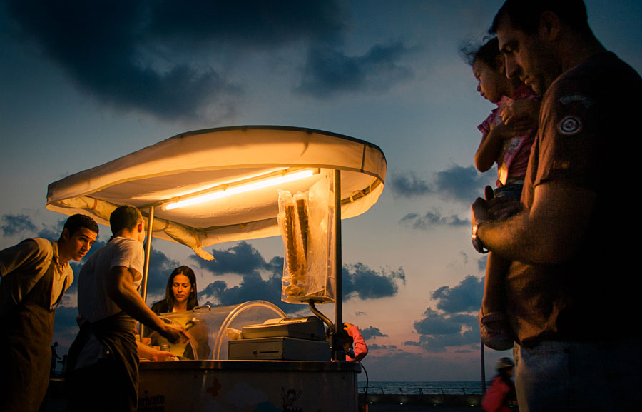 People gathered around a lit street food cart at sunset with dramatic clouds