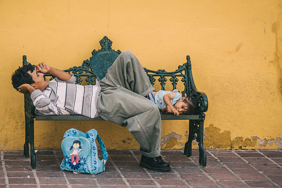 Father and son asleep on an ornate iron bench against a yellow wall in Latin America