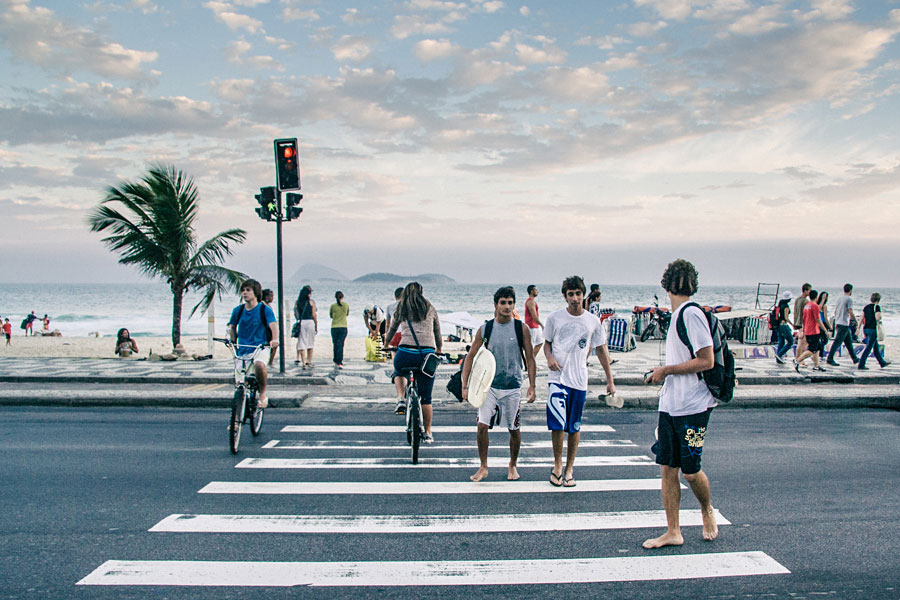Surfers and cyclists crossing a beachside road with ocean and mountains in the background