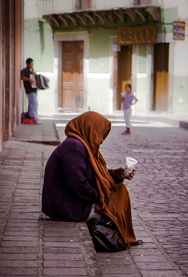 Elderly woman wrapped in a brown shawl sitting on a cobblestone street in Mexico