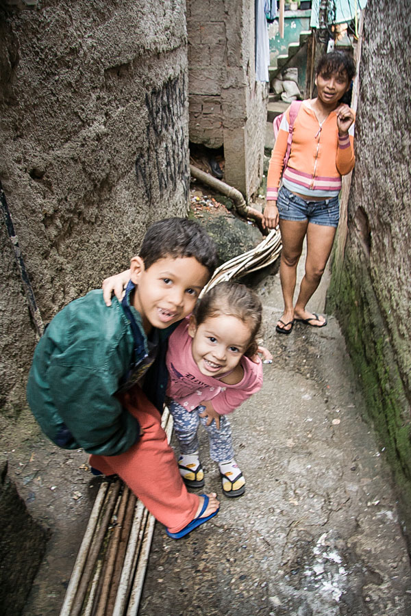 Two children grinning in a narrow favela alleyway with a woman standing behind them