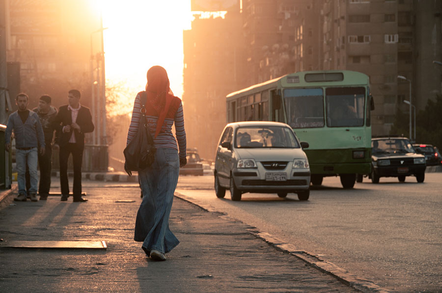 Woman silhouetted walking across an urban street at golden hour with a bus passing behind