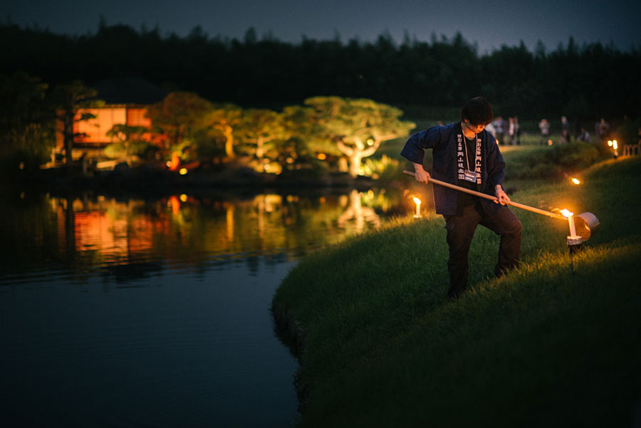 Worker lighting torches along a Japanese garden pond at night with illuminated trees reflecting in water