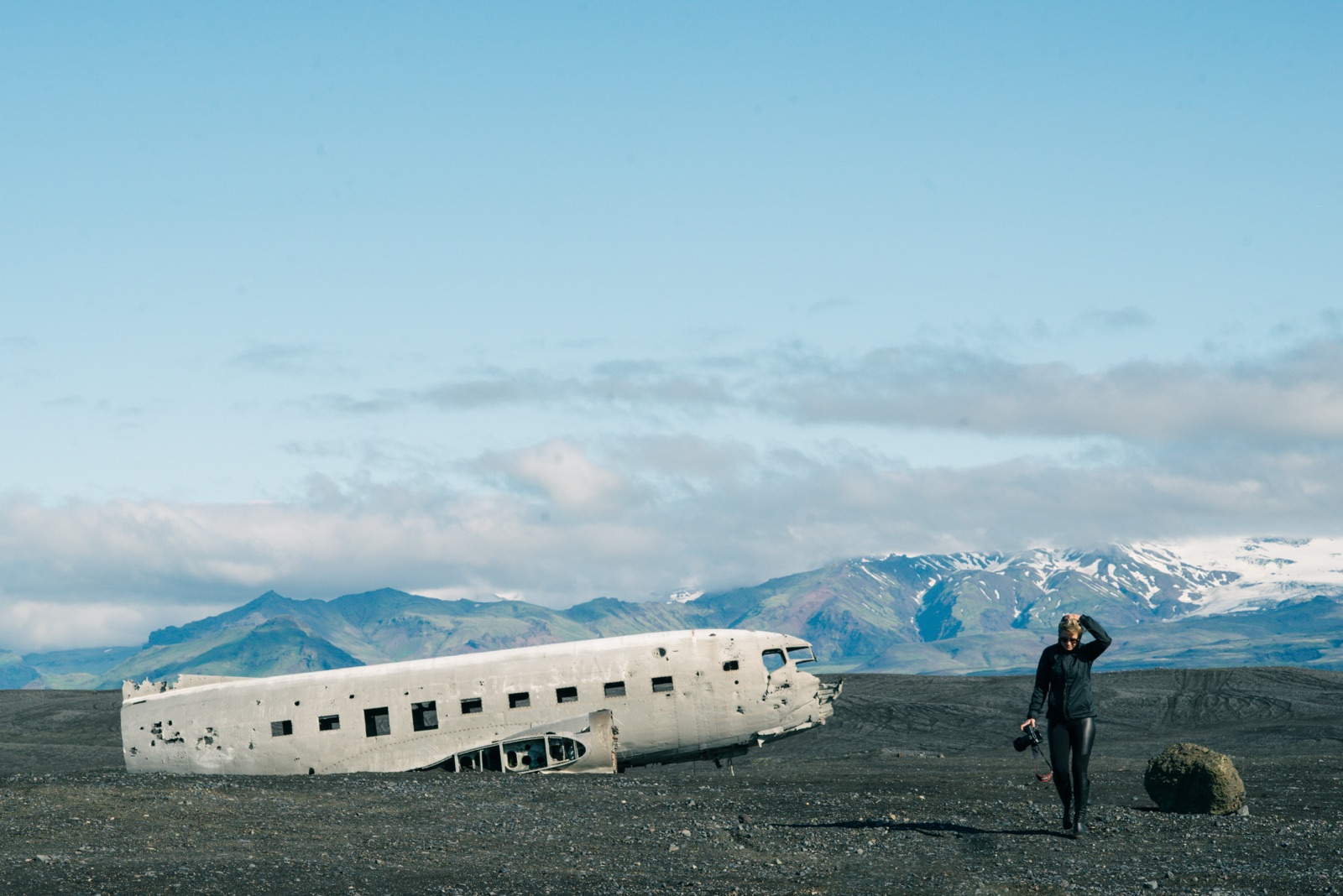 Woman in black walking from the Sólheimasandur DC-3 plane wreck on black sand, Iceland — snow-capped mountains behind