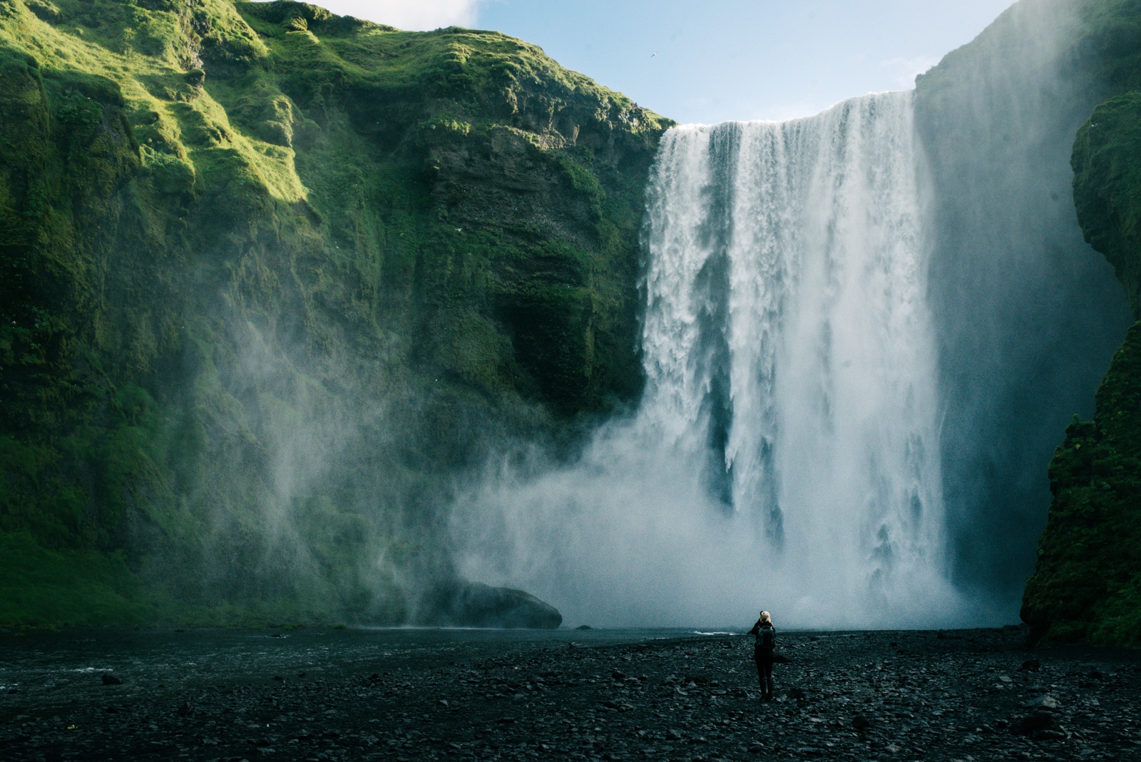 Skógafoss waterfall in Iceland with a single figure at the base for scale, moss-covered cliff above