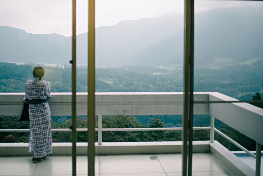 Person in a yukata looking out from a balcony at misty mountains through glass doors