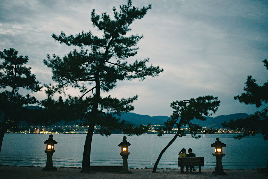 Couple sitting on a bench by the water at dusk under pine trees with stone lanterns glowing