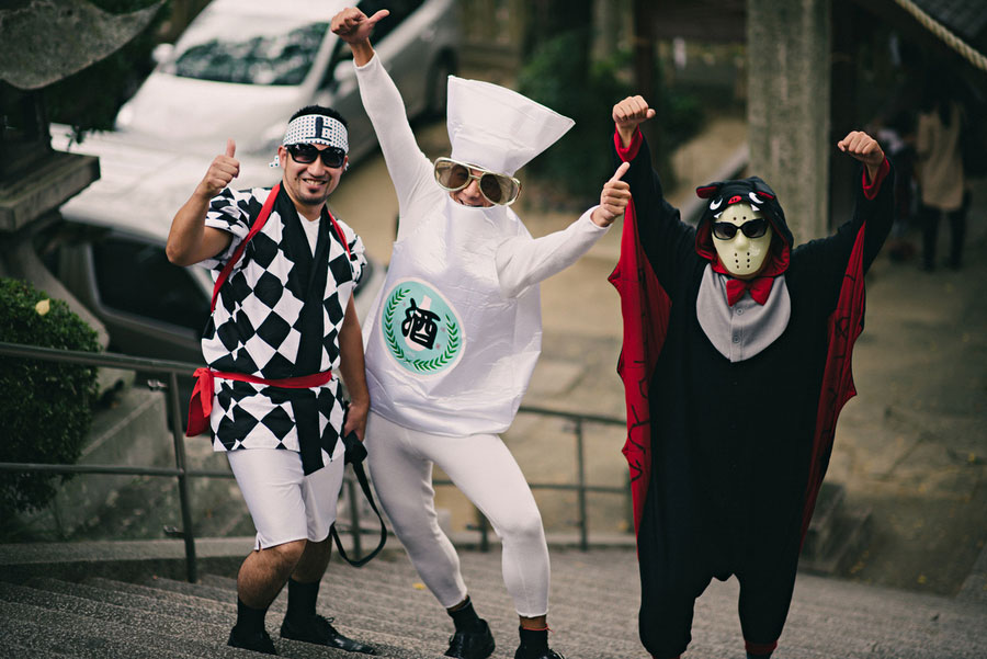 Three men in costume posing on stone steps with checkered pattern, sake bottle, and vampire cape