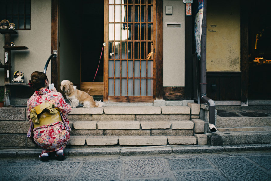Woman in a floral kimono crouching to pet a dog resting on steps of a traditional shopfront