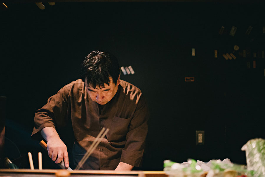 Japanese chef focused on preparing food behind a counter in a dimly lit restaurant kitchen