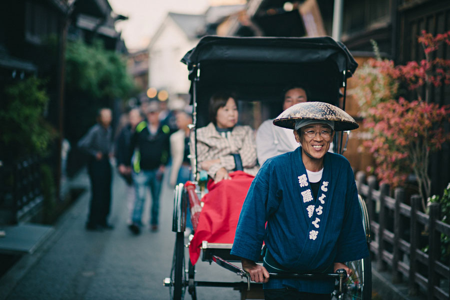 Rickshaw driver in a straw hat smiling while pulling passengers through a narrow historic street