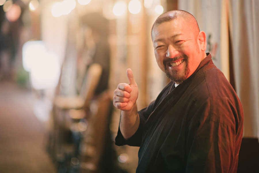 Bearded man in dark clothing giving a thumbs up and wide smile outside a Japanese bar at night